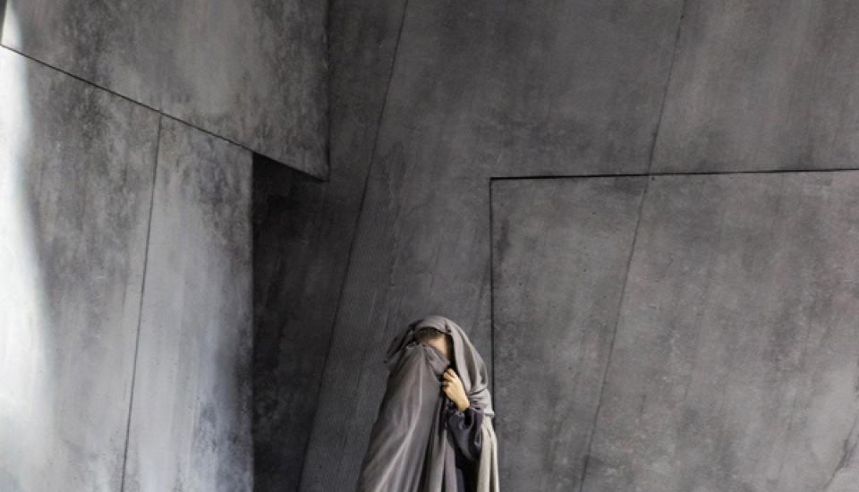 Un personnage avec une cape grise se cache le visage dans un décor gris. Au premier plan, des pierres, des branchages et peut-être une boîte.